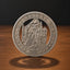 A round silver-toned commemorative coin featuring a grim reaper design, standing upright on a wooden surface against a dark background.
