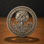 A circular silver-toned commemorative coin or medallion stands upright on a wooden surface against a dark background. The coin features an embossed figure in flowing robes holding a sword, surrounded by stars and the text 'E PLURIBUS UNUM' and '1921'.