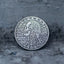 Close-up of a coin engraved with a skull surrounded by flowing baroque curly hair and scrollwork, spine detail below, Liberty above, In God We Trust, dated 1964