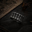 Close-up of a wide silver ring featuring crossed bone and spine detailing with oxidized black finish, resting on a dark stone surface beside aged parchment