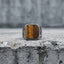 Tiger eye square signet ring standing upright on a concrete ledge with a grey cement wall background, showing the full amber stone face and floral scroll engraving on both band shoulders in natural diffused light
