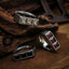 Three square gemstone signet rings arranged on a dark wood and rusted metal surface, showing white crystal, red garnet, and black onyx stone variants with engraved silver bands and dried leaves in the background