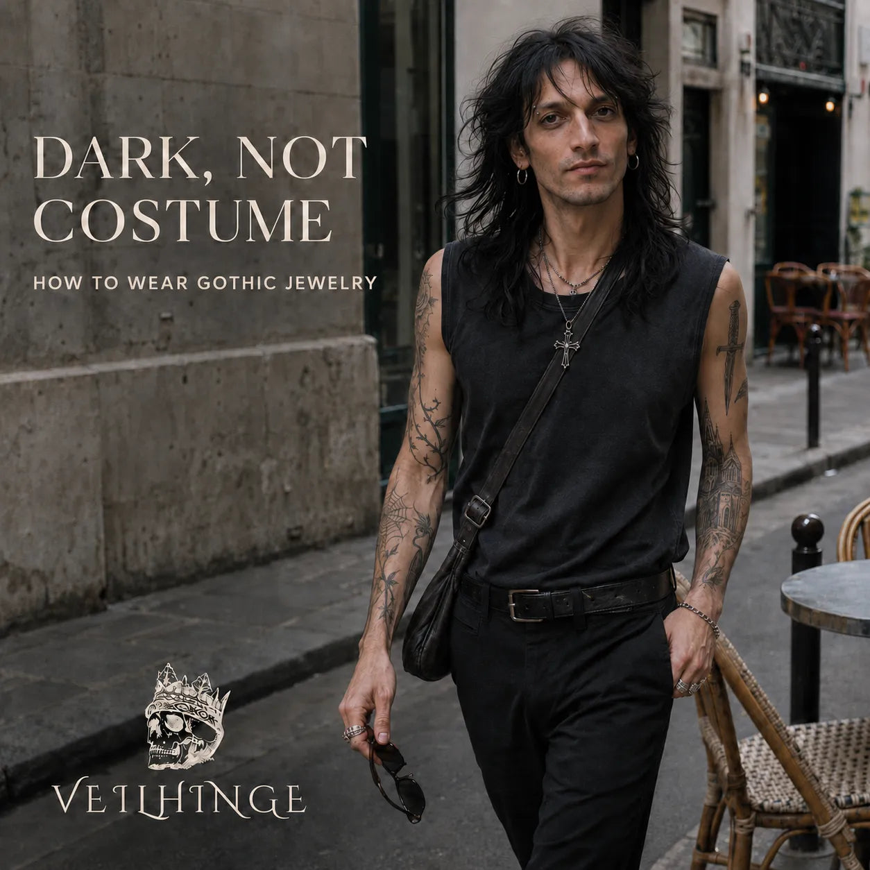 Tattooed man wearing a gothic cross pendant and rings with a restrained black outfit on a city street.