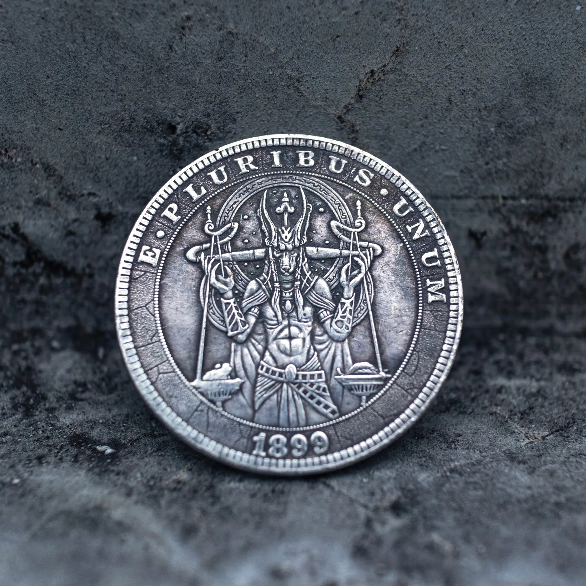 A round silver-toned coin or medallion featuring a detailed central figure and the text 'E PLURIBUS UNUM' around the edge, with '1899' at the bottom, photographed on a dark textured surface.