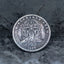 A round silver-toned coin or medallion featuring a detailed central figure and the text 'E PLURIBUS UNUM' around the edge, with '1899' at the bottom, photographed on a dark textured surface.