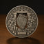 A round silver-toned medallion or coin-like collectible with an embossed classical design, featuring a central shield motif, two human figures in relief, and the text 'E PLURIBUS UNUM' around the edge, photographed standing on a wooden surface against a dark background.