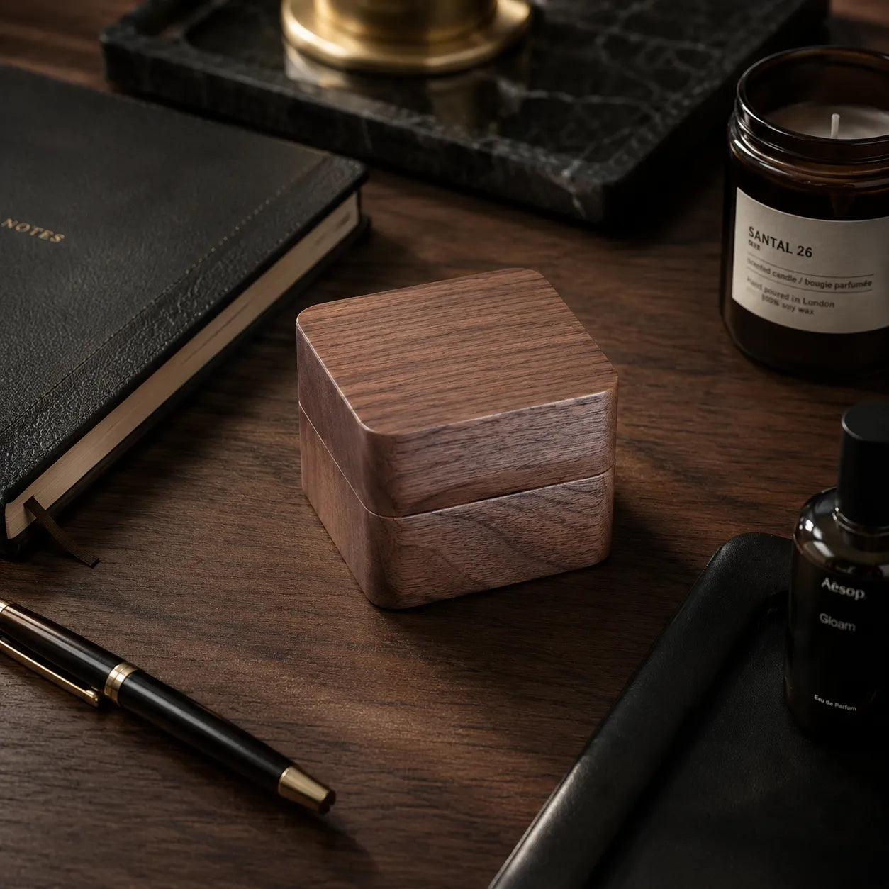 A small square wooden keepsake box with a visible wood grain finish sits on a dark wooden desk surrounded by lifestyle props including a black leather notebook, a pen, a candle, and a bottle.