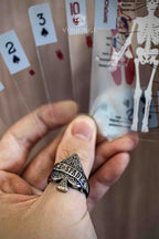 Ace of Spades ring worn while holding a fan of spade playing cards, baroque-engraved spade face ring with gothic skull poster in the background