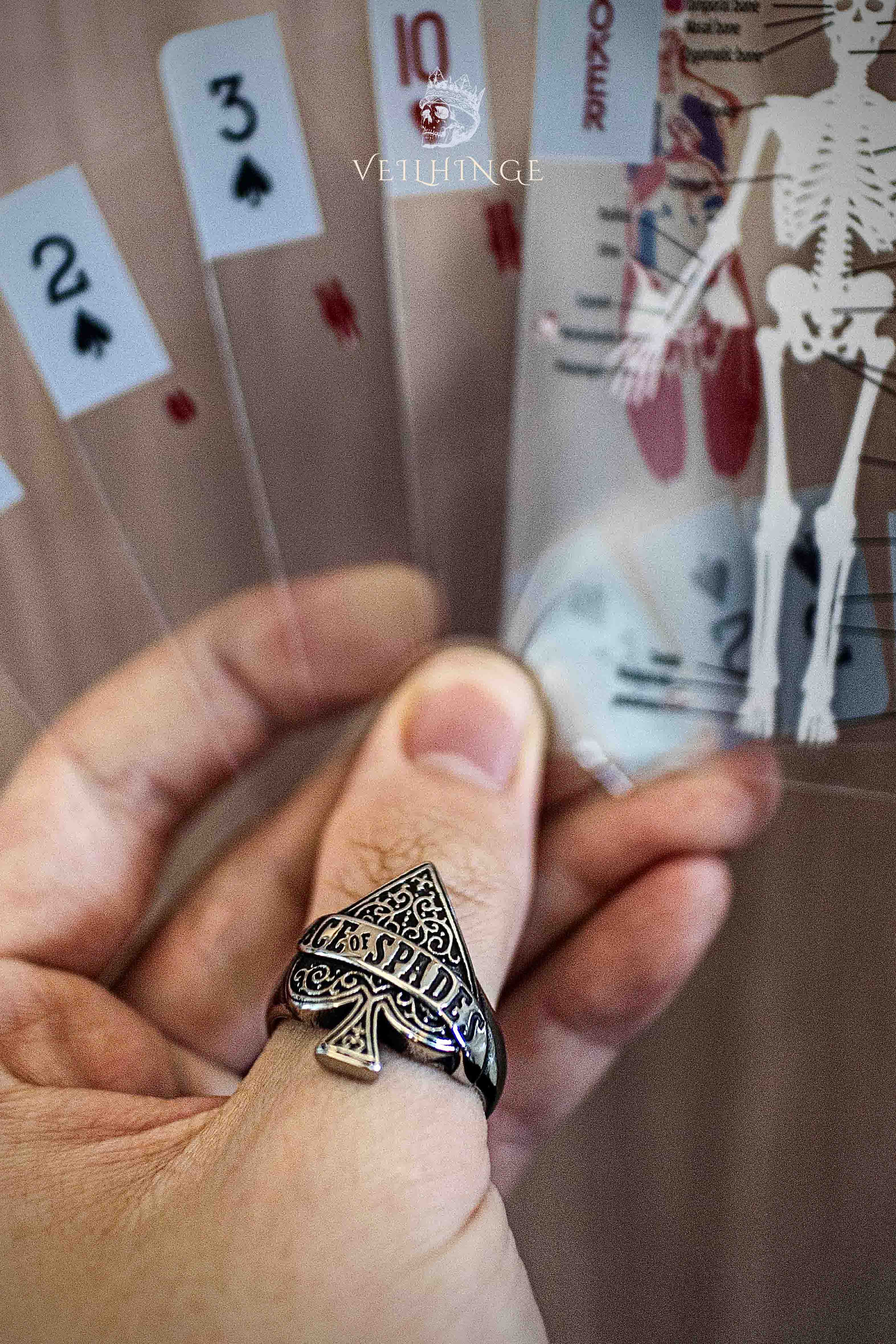 Ace of Spades ring worn while holding a fan of spade playing cards, baroque-engraved spade face ring with gothic skull poster in the background
