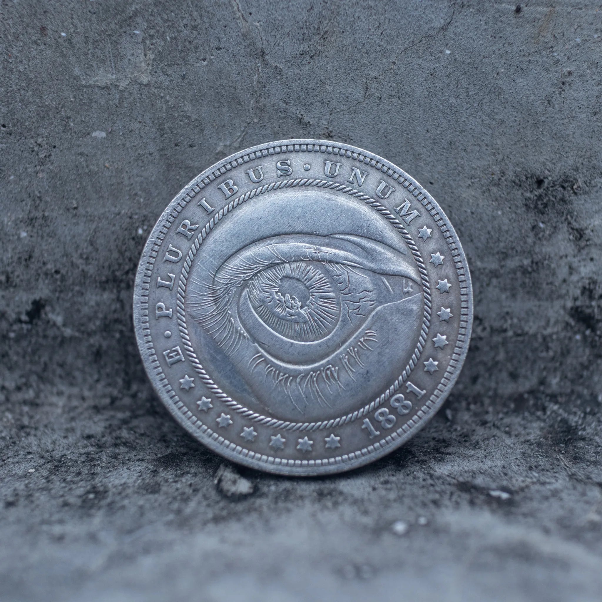 Close-up of a coin engraved with a single large eye, detailed iris and pupil, concentric ripple rings radiating outward, E Pluribus Unum border, dated 1881, on dark stone