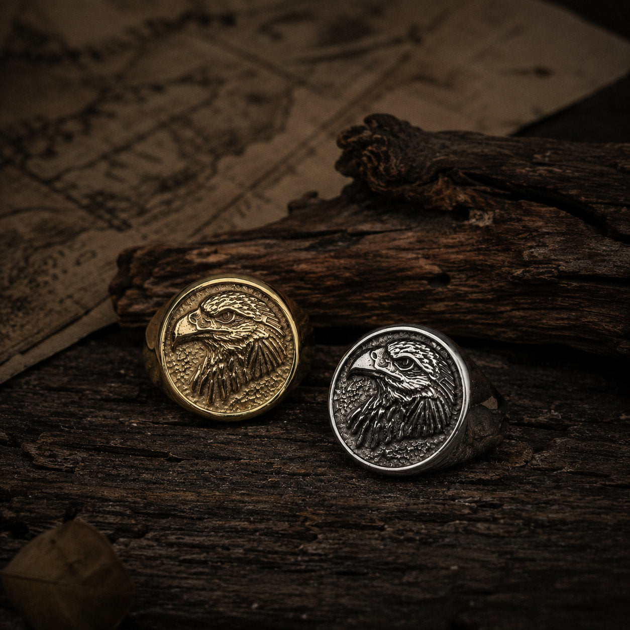 Gold and silver bald eagle signet rings placed side by side on a wood surface with both engraved faces visible, photographed in warm natural light showing the color contrast between the two finishes