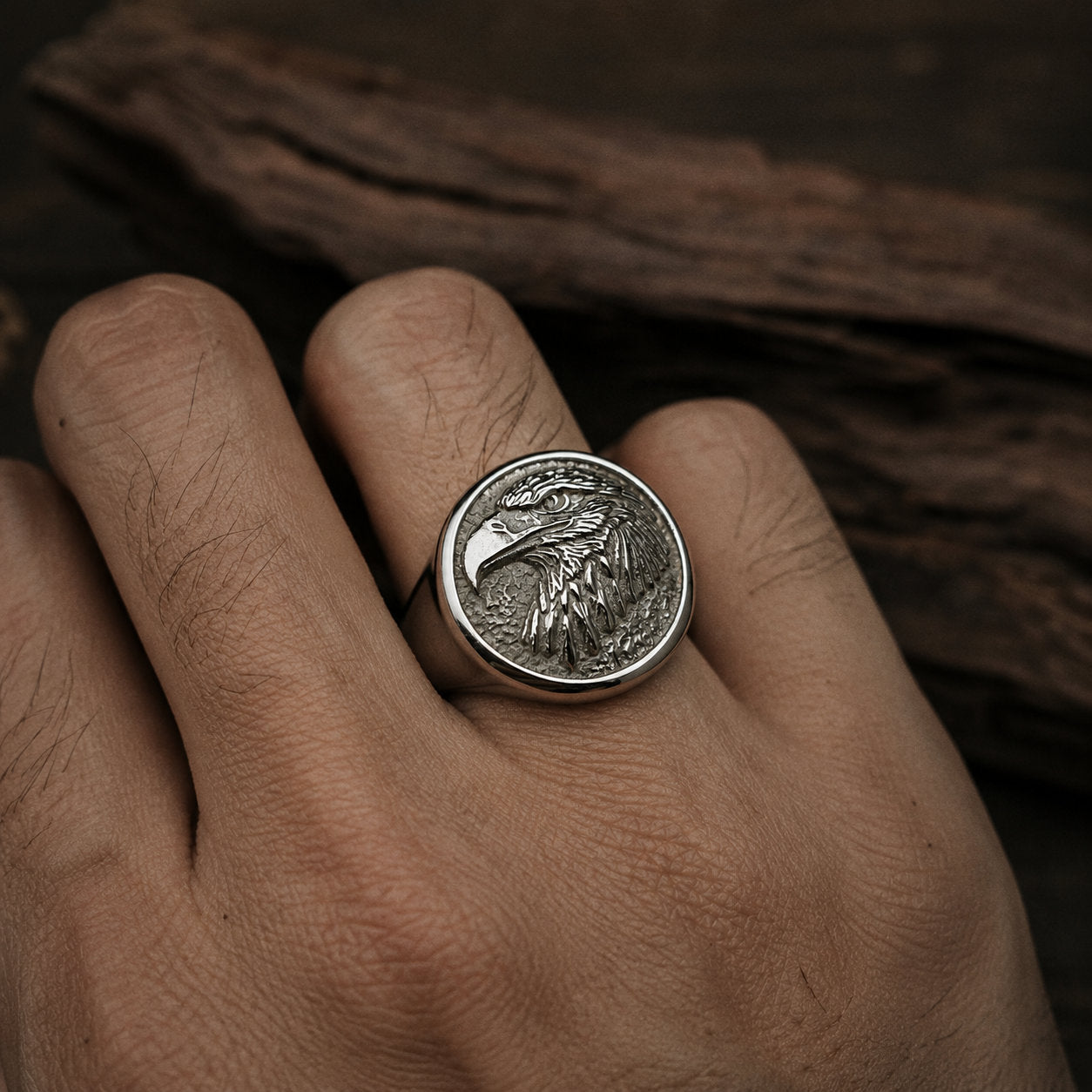 Man's hand wearing an antique silver bald eagle signet ring on the index finger, photographed against a dark wood surface in side lighting