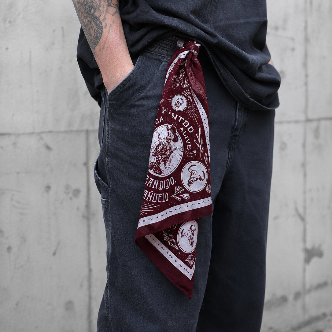 Burgundy Bandido bandana folded and tucked into the side pocket of grey trousers, with the skull cowboy graphic facing outward, worn by a tattooed man against a concrete wall