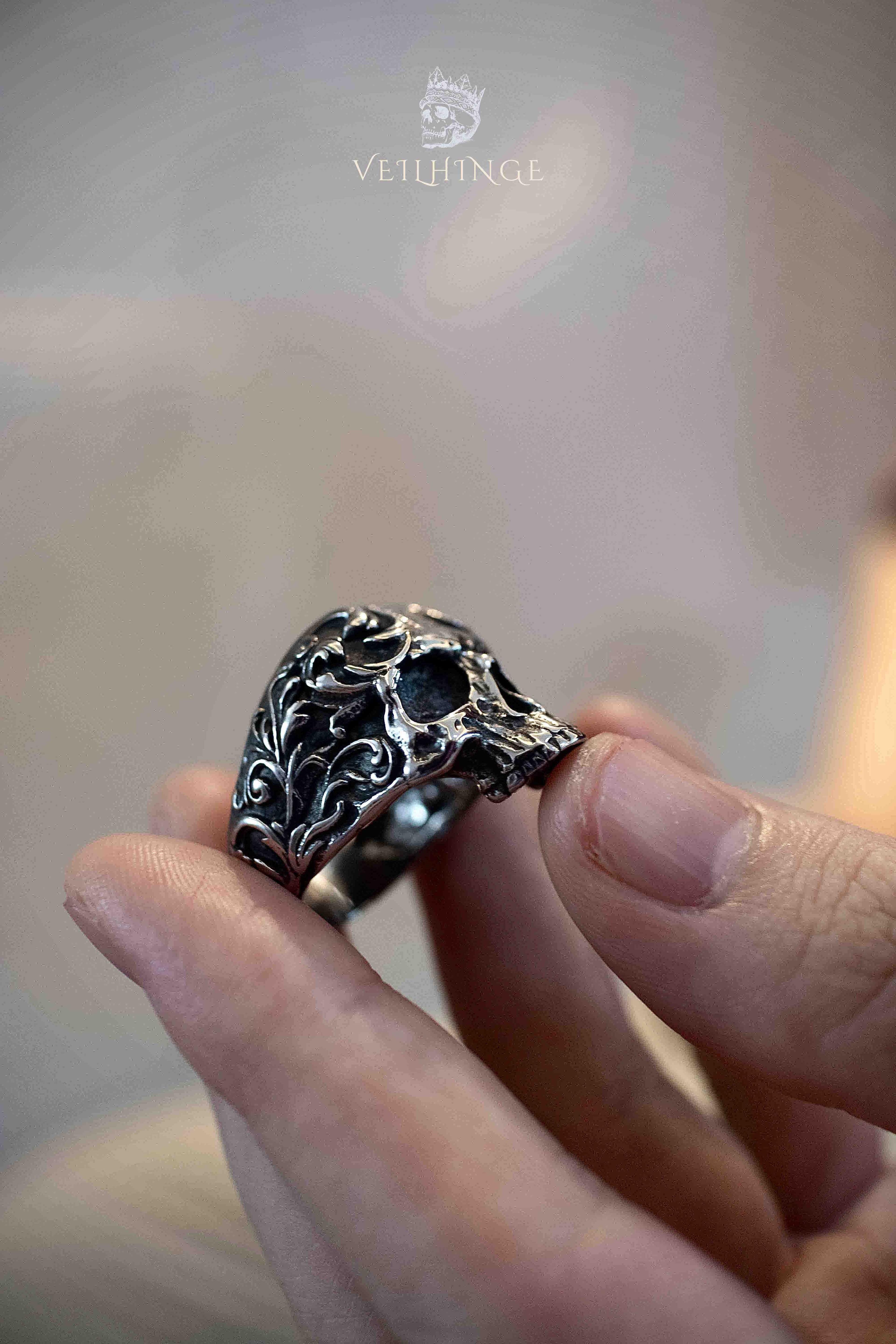 Baroque floral skull ring held between fingers showing the full face, oxidized silver with scrollwork vines covering the cheekbones and forehead of the skull