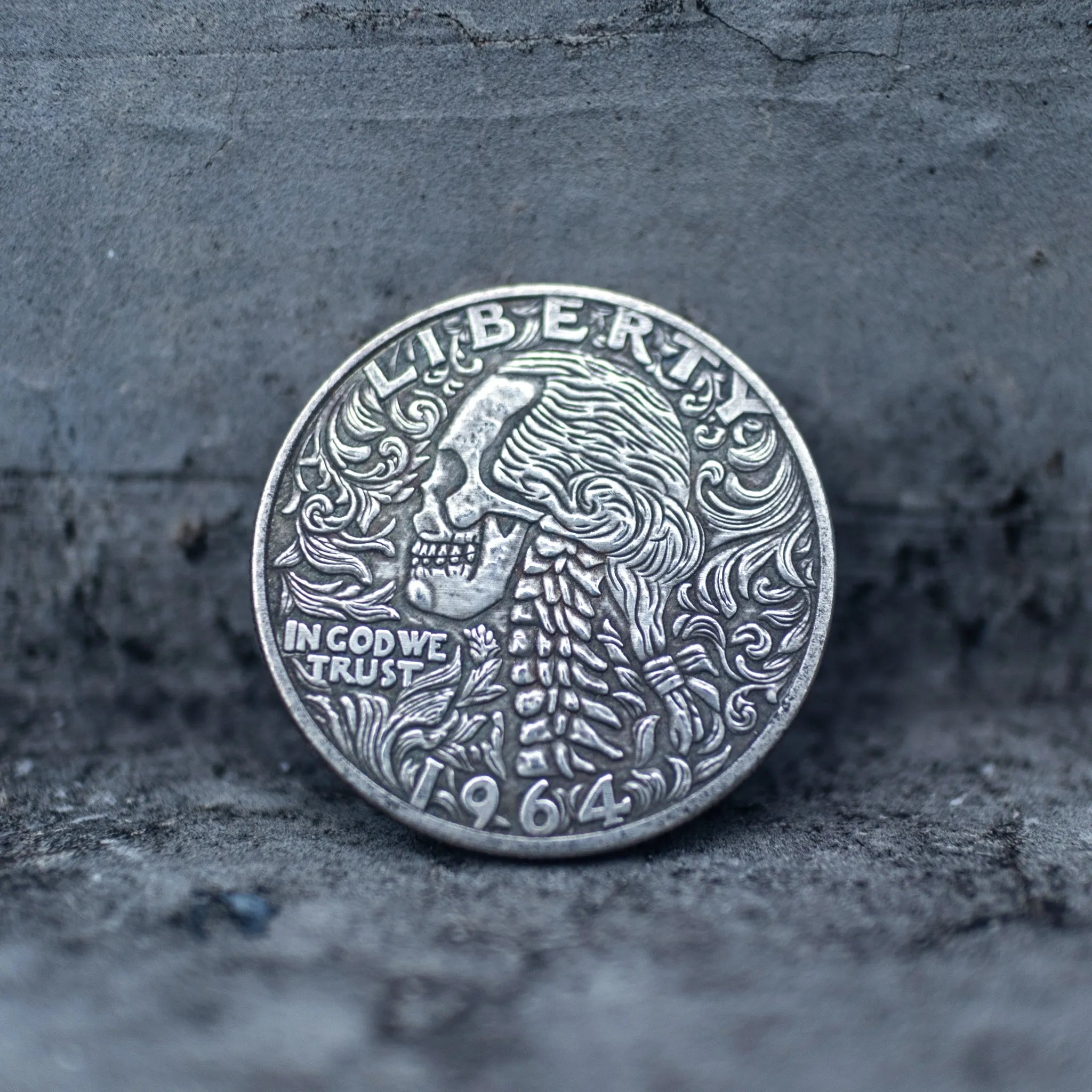 Close-up of a coin engraved with a skull surrounded by flowing baroque curly hair and scrollwork, spine detail below, Liberty above, In God We Trust, dated 1964