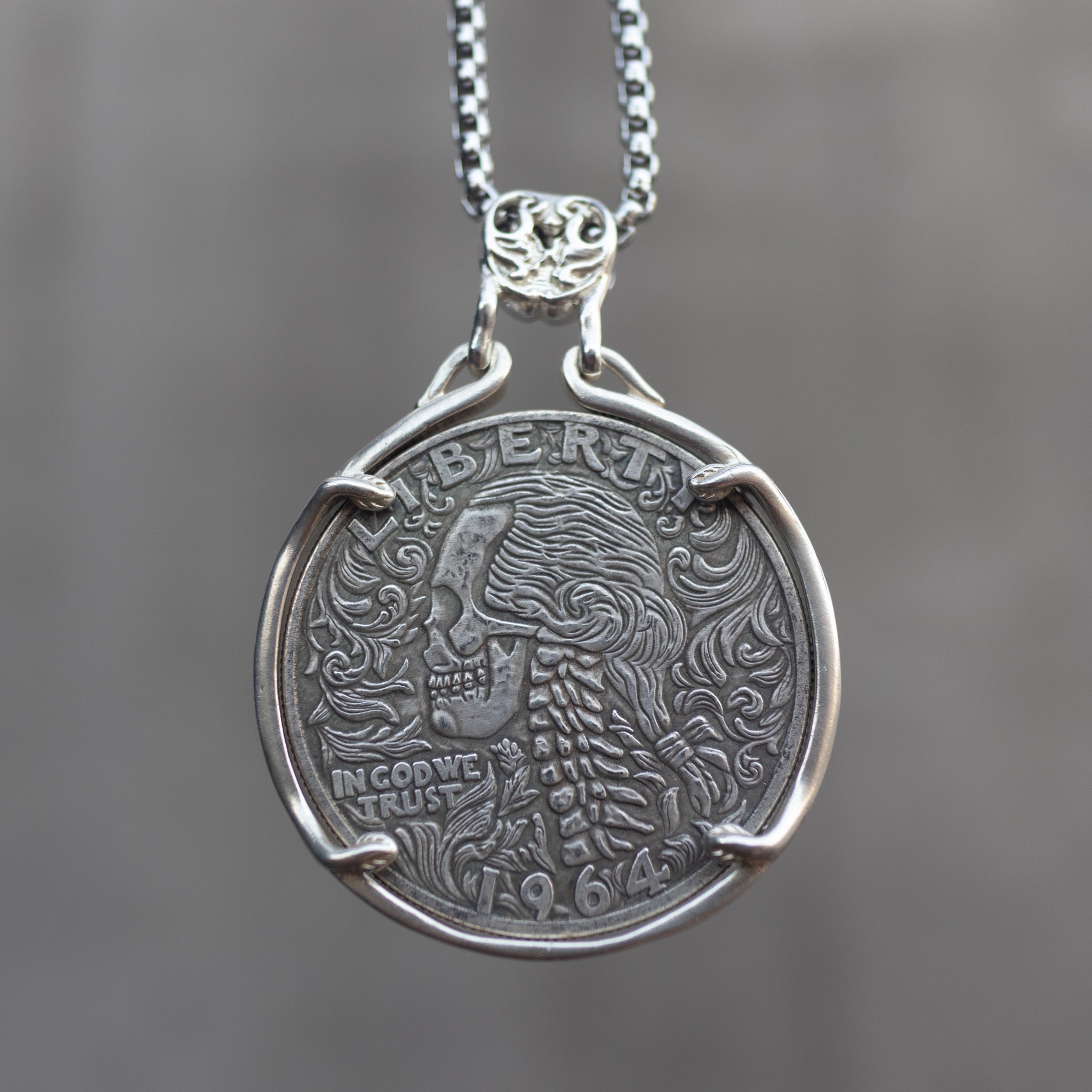 Coin pendant engraved with a baroque skull with flowing curly hair and scrollwork details, spine below, Liberty inscription, dated 1964, in a four-prong bezel on a chain