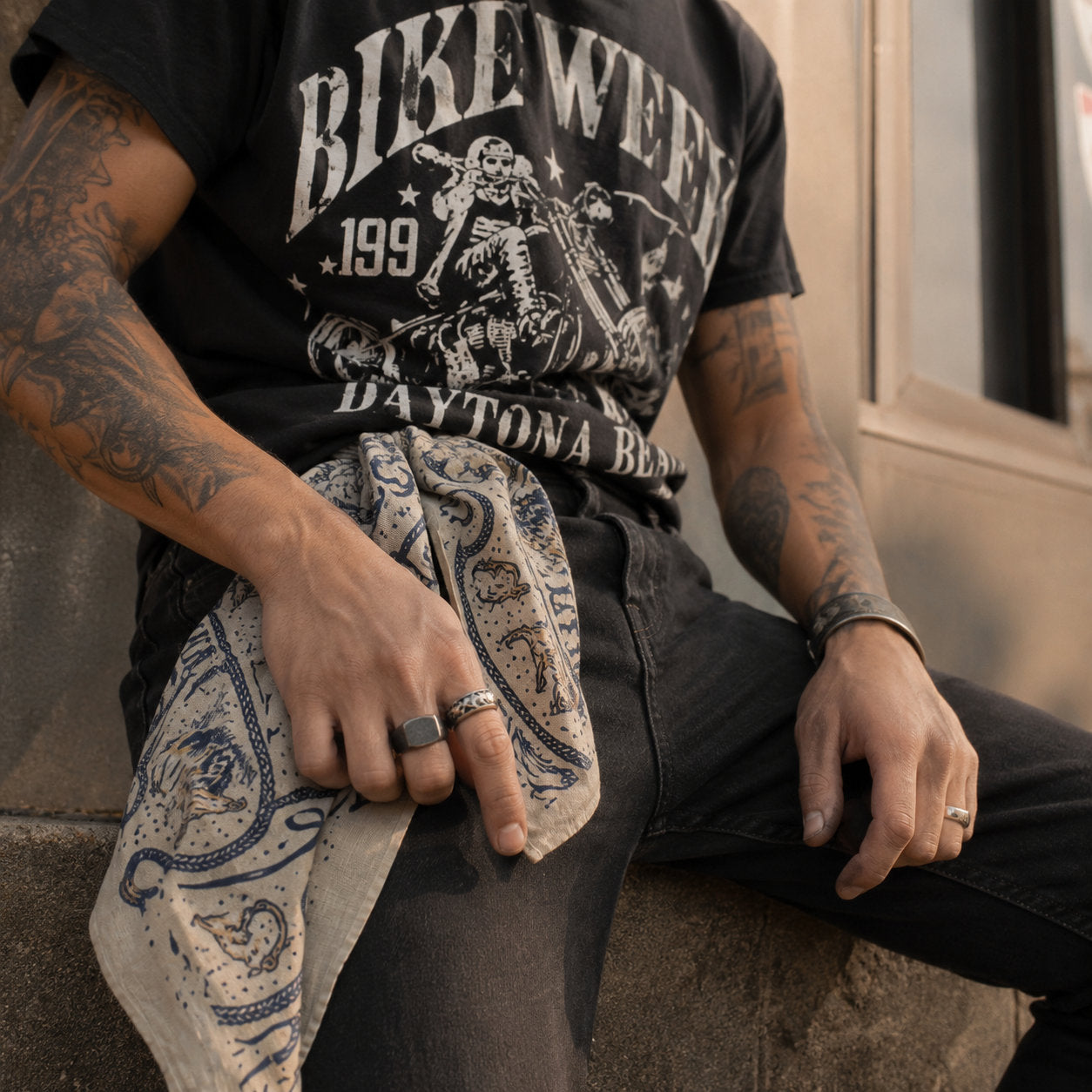 White and navy western bandana folded and tucked into the waistband of dark jeans, worn with a black Bike Week Daytona Beach graphic tee by a tattooed man with rings on his hands