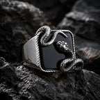 Close-up of a black onyx signet ring on dark rock showing a silver serpent coiled around the square stone with the snake head raised at the upper corner and detailed scale texture visible