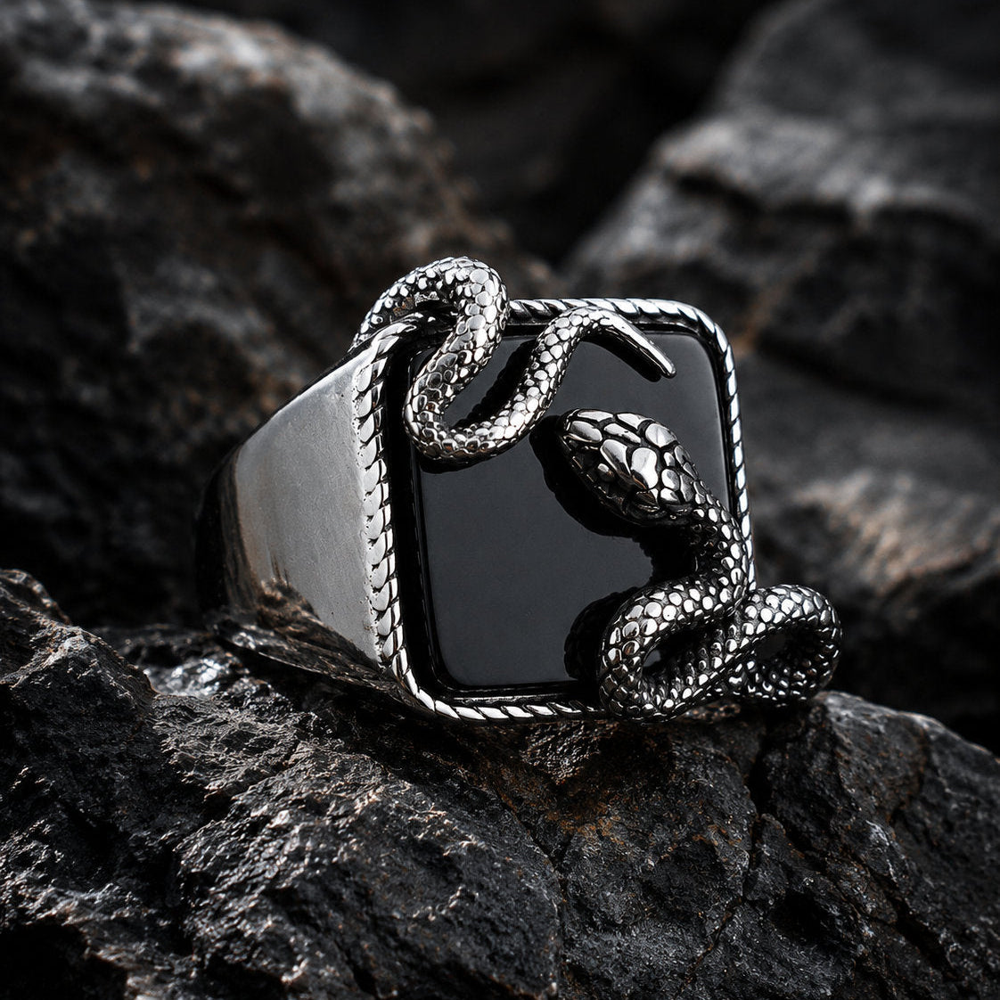 Close-up of a black onyx signet ring on dark rock showing a silver serpent coiled around the square stone with the snake head raised at the upper corner and detailed scale texture visible