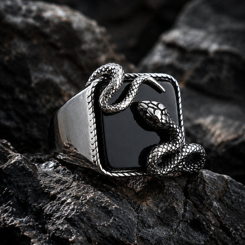 Close-up of a black onyx signet ring on dark rock showing a silver serpent coiled around the square stone with the snake head raised at the upper corner and detailed scale texture visible