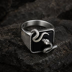Black onyx snake signet ring photographed from a side-rear angle on dark rock, showing the contrast between the smooth polished silver band and the textured serpent sculpture on the stone face