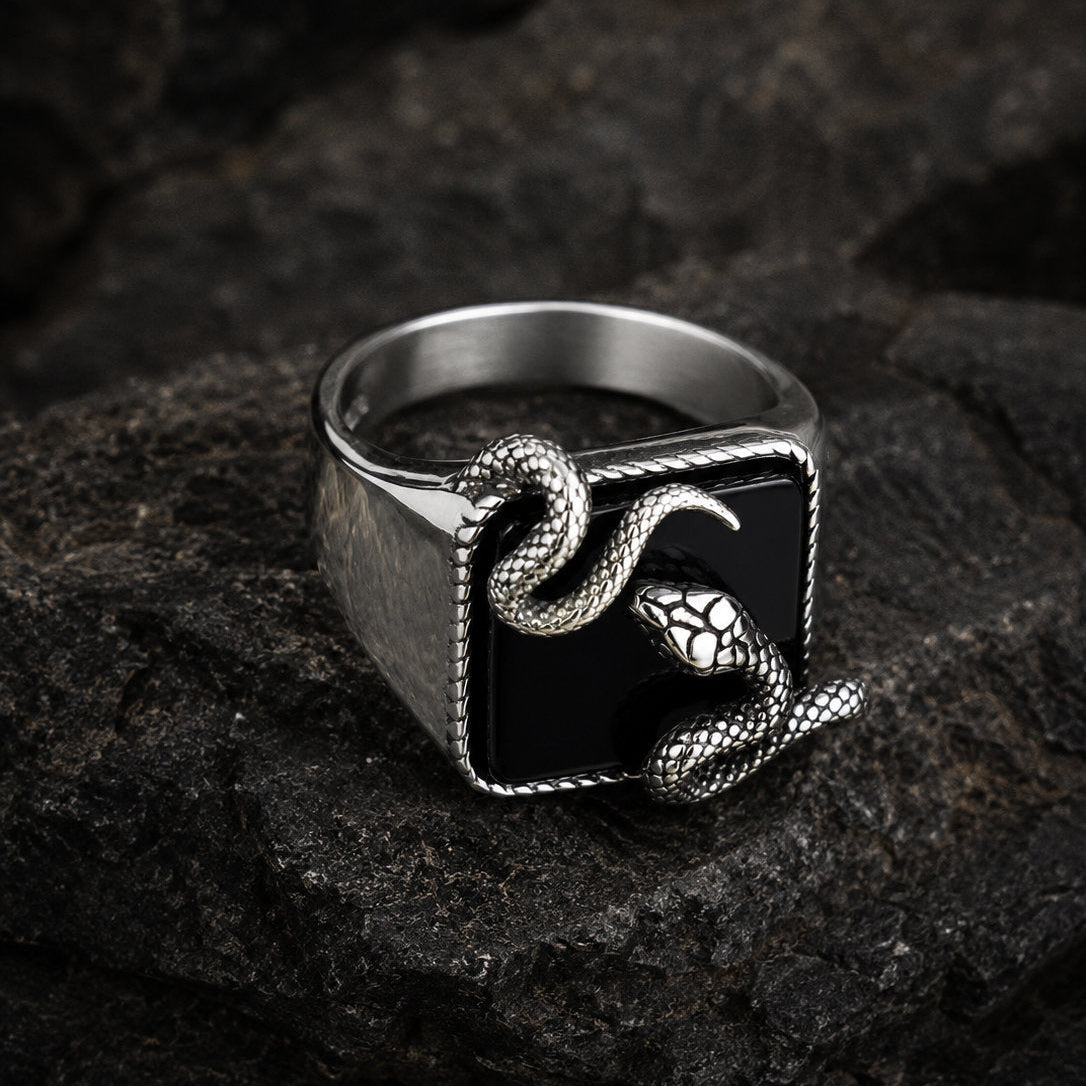 Black onyx snake signet ring photographed from a side-rear angle on dark rock, showing the contrast between the smooth polished silver band and the textured serpent sculpture on the stone face