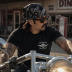 Man wearing a black skull-print bandana on his head while riding a motorcycle, shot in front of a vintage roadside diner