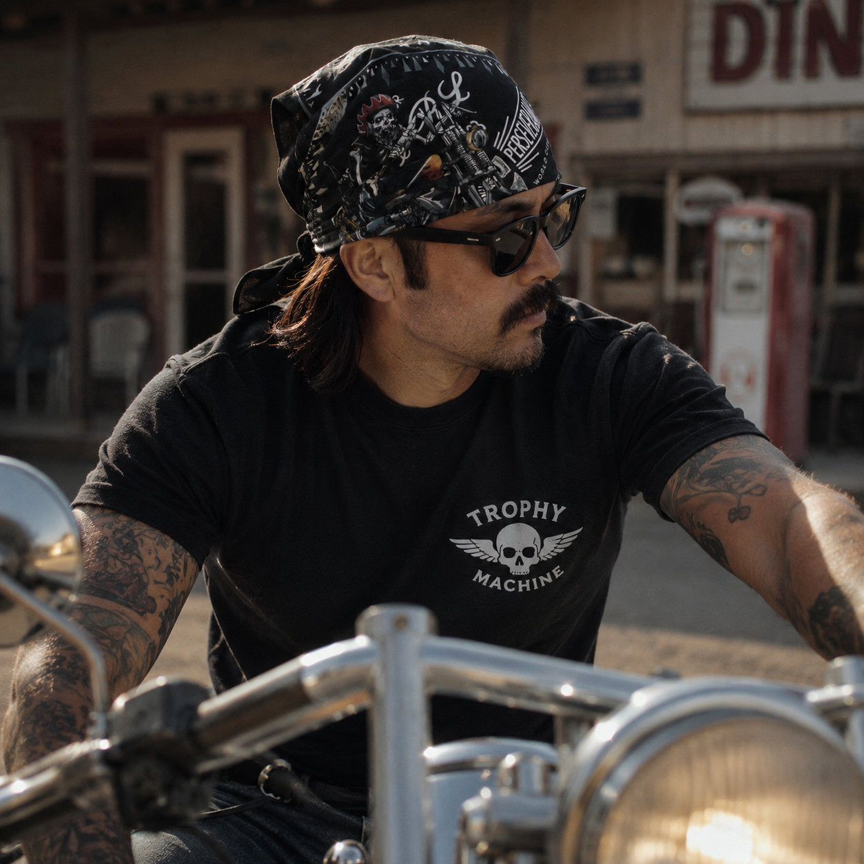 Man wearing a black skull-print bandana on his head while riding a motorcycle, shot in front of a vintage roadside diner