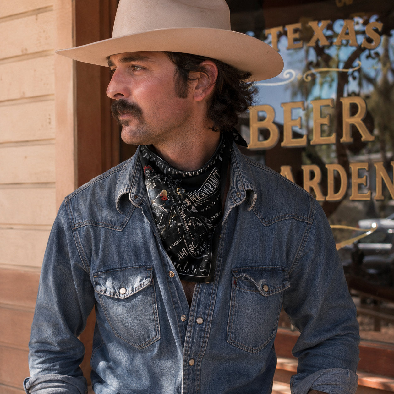 Man wearing a black printed bandana tied around his neck over a denim shirt, styled in a western bar setting