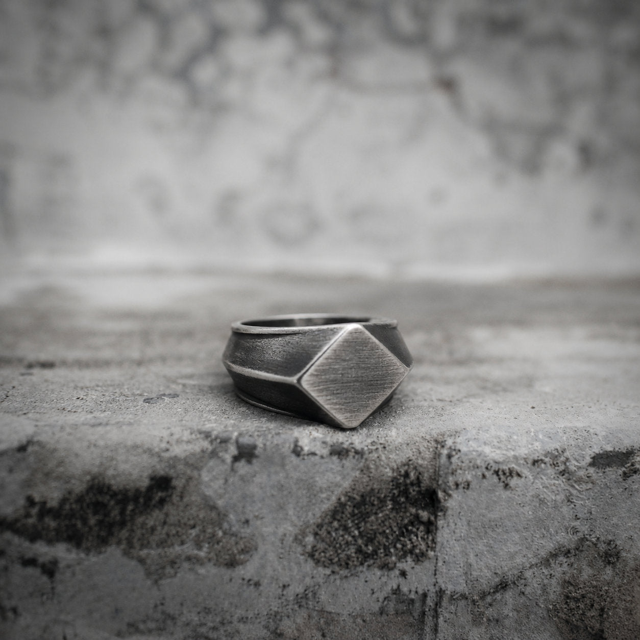 Oxidized silver square signet ring resting on a concrete surface, showing the geometric diamond-cut face with brushed matte texture and angular beveled edges in close-up detail Main image