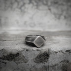 Oxidized silver square signet ring resting on a concrete surface, showing the geometric diamond-cut face with brushed matte texture and angular beveled edges in close-up detail