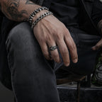 Tattooed man in an all-black biker outfit sitting with his hand resting on his knee, wearing a square signet ring on his index finger alongside a Cuban link bracelet and chain details