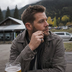 Man in a grey washed leather jacket resting his chin on his hand, wearing a small square signet ring on his index finger, photographed outdoors with a mountain backdrop