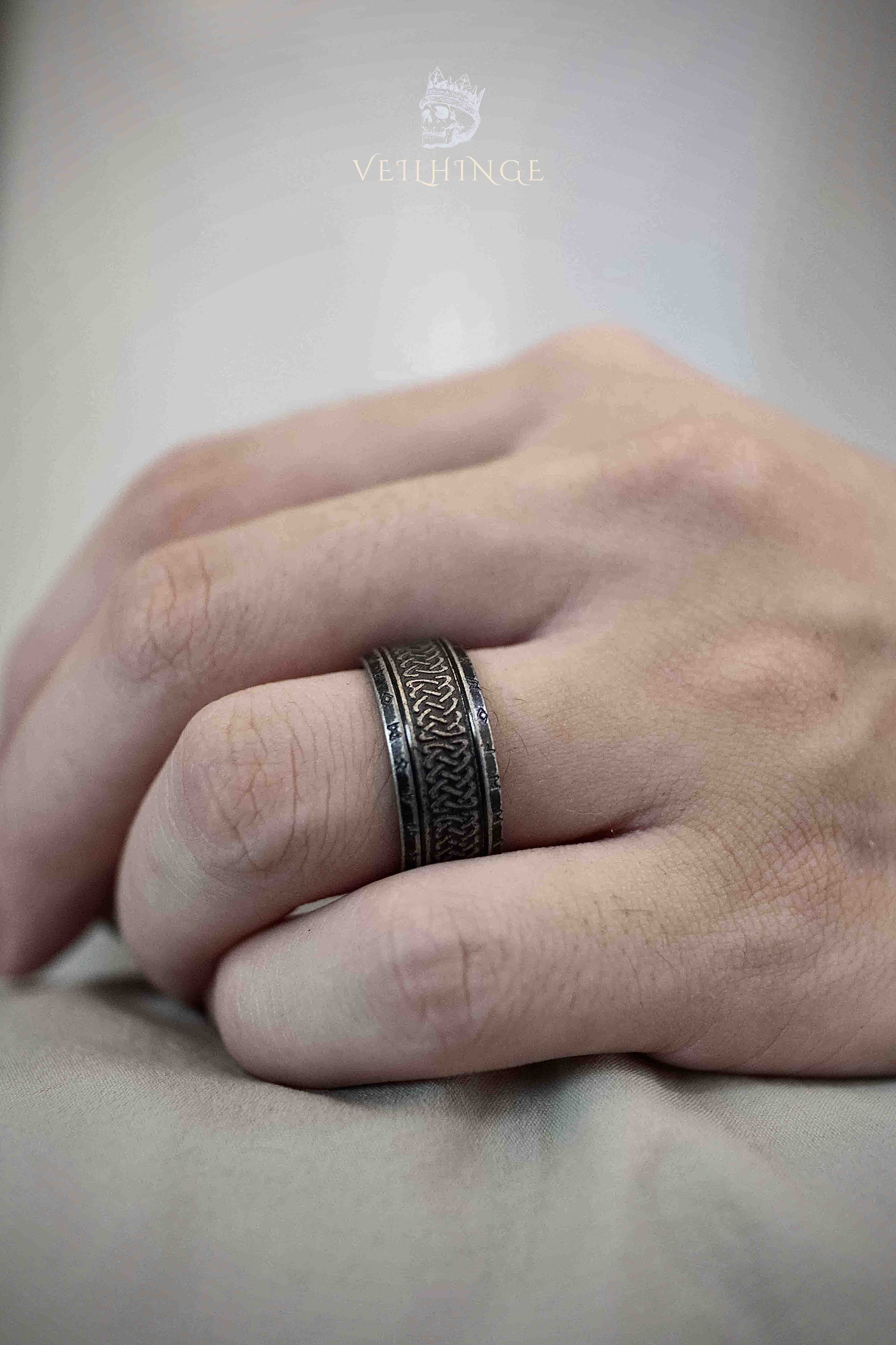 Wide Celtic knotwork band ring worn on a middle finger, dense interlaced pattern covering the full band width in deeply oxidized dark metal, hands resting together