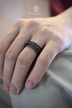 Black oxidized wide band ring with a rotating chain layer set into the center, worn on a ring finger resting on fabric, beaded edges framing the spinning chain