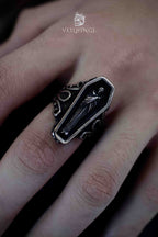 Coffin-shaped signet ring with black enamel face featuring a cloaked skeleton lying in repose, silver baroque scrollwork on the shoulders, worn on a hand