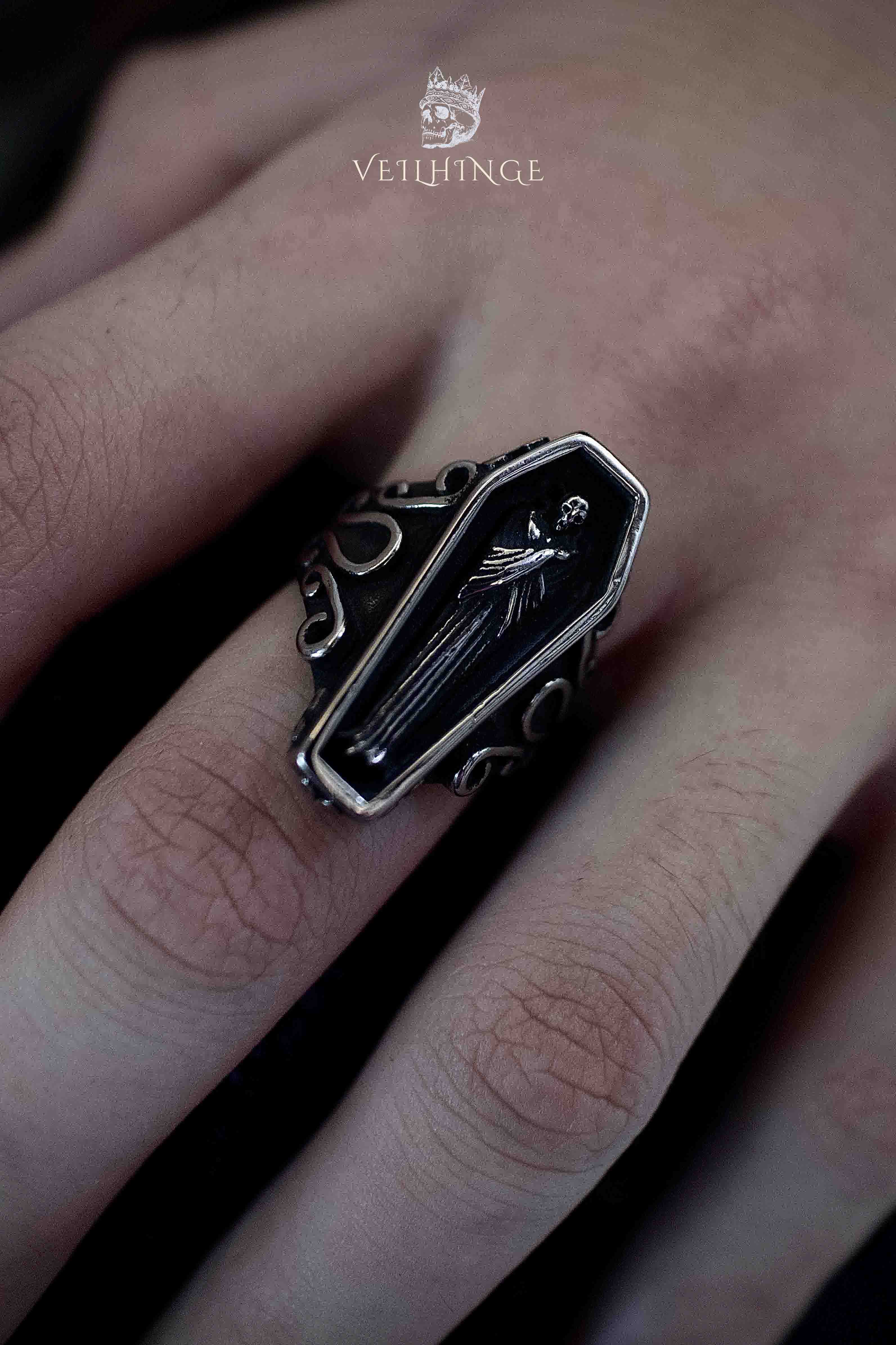 Coffin-shaped signet ring with black enamel face featuring a cloaked skeleton lying in repose, silver baroque scrollwork on the shoulders, worn on a hand