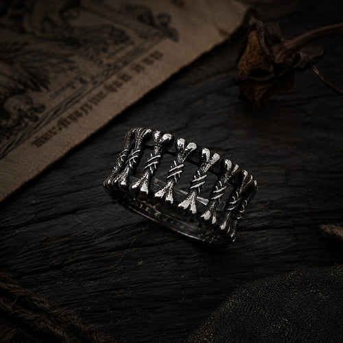 Close-up of a wide silver ring featuring crossed bone and spine detailing with oxidized black finish, resting on a dark stone surface beside aged parchment