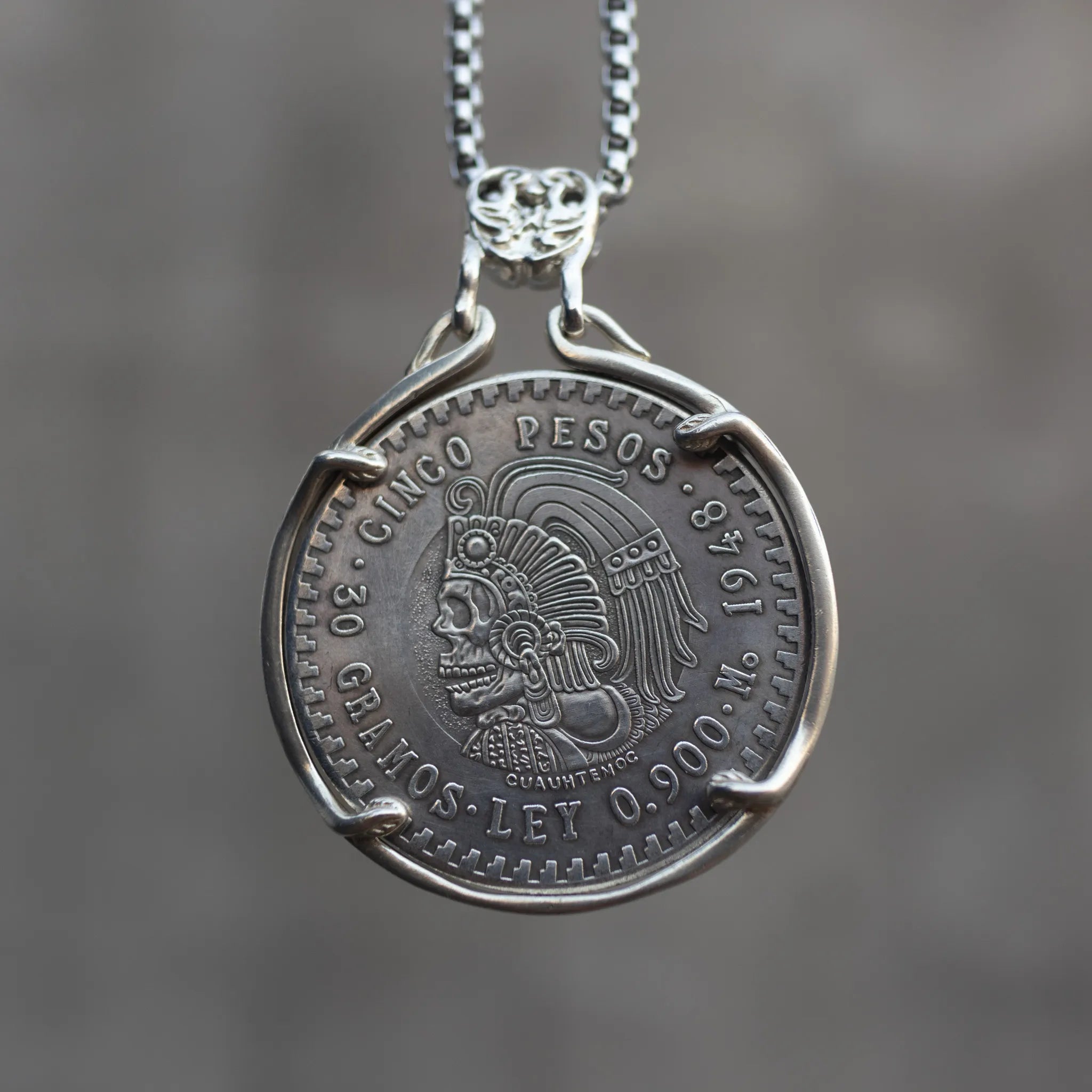 Cinco Pesos coin pendant featuring a skull-faced Cuauhtémoc in Aztec headdress, set in a four-prong bezel with skull bail on a box chain
