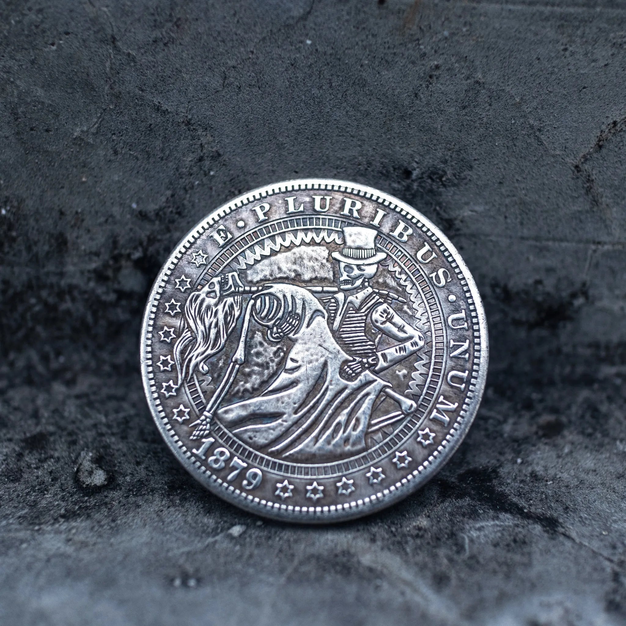 Close-up of a coin engraved with two skeletons dancing — male skeleton in top hat and suit leading a female skeleton in a long dress bending backward, E Pluribus Unum border, dated 1879