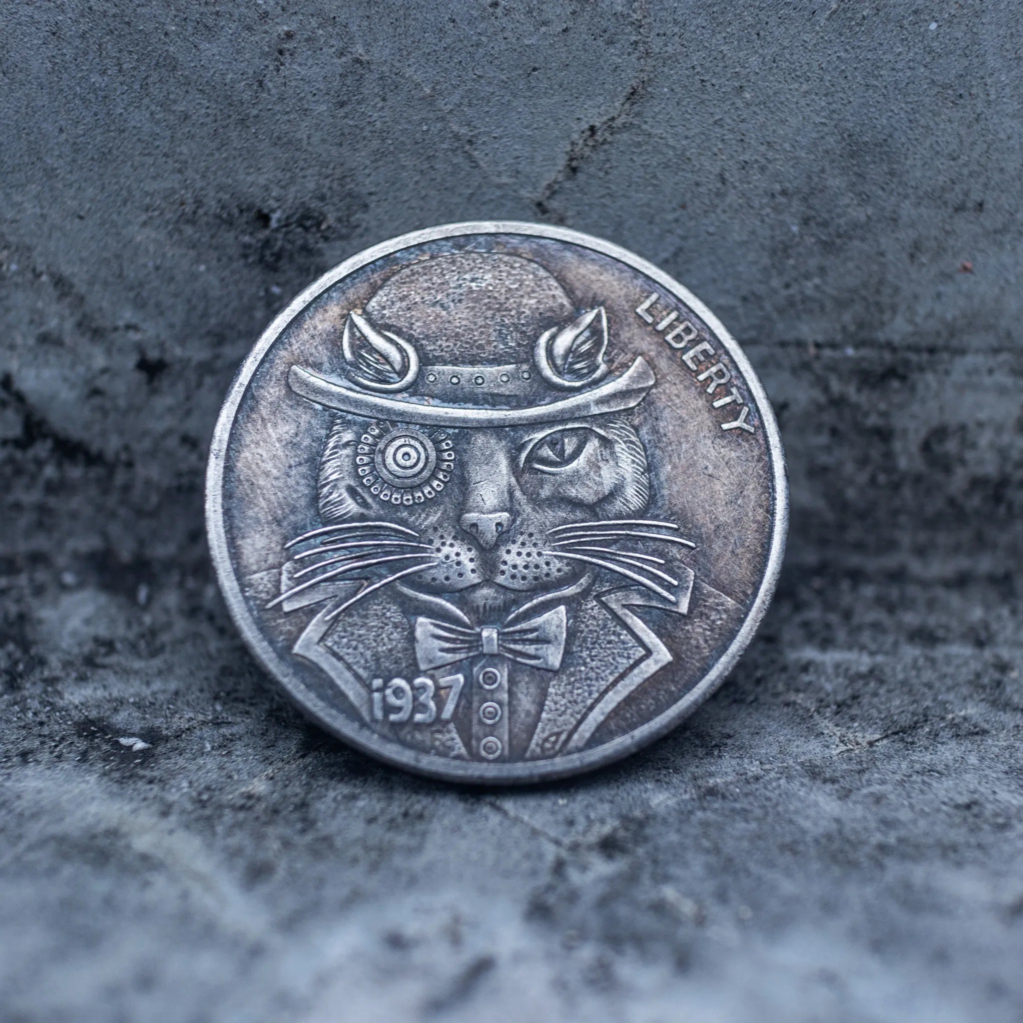 Close-up of a coin engraved with a dapper cat wearing a bowler hat and bow tie, concentric circle eyes, whiskers, Liberty inscription, dated 1937