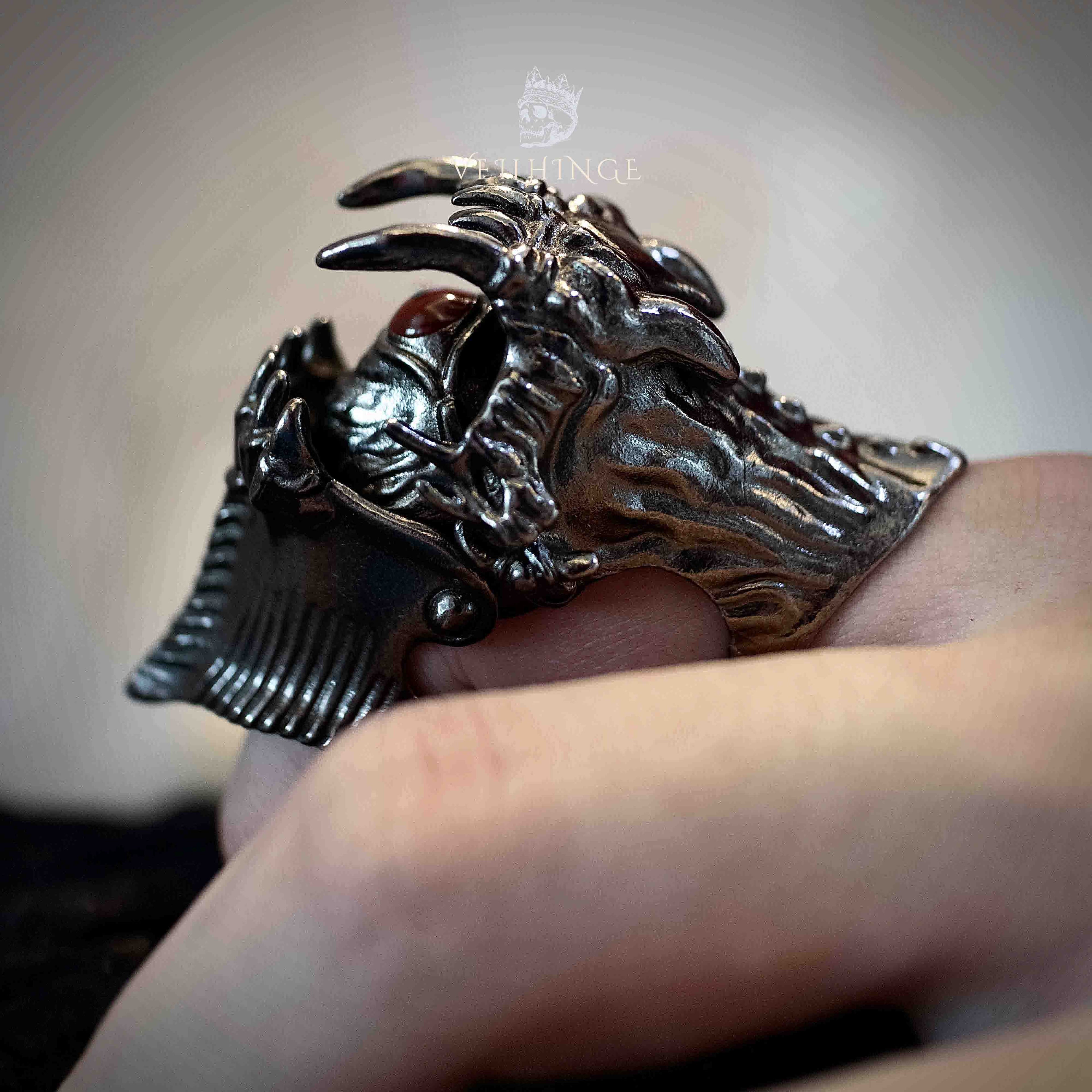 Demon mouth armor ring on a bent finger with the jaw open, two sharp fangs visible at the top and the eye revealed inside the open mouth, dark oxidized metal