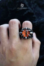 Dragon claw ring gripping an orange faceted stone, with cloaked skull figures on each shoulder, worn on a hand against a black background