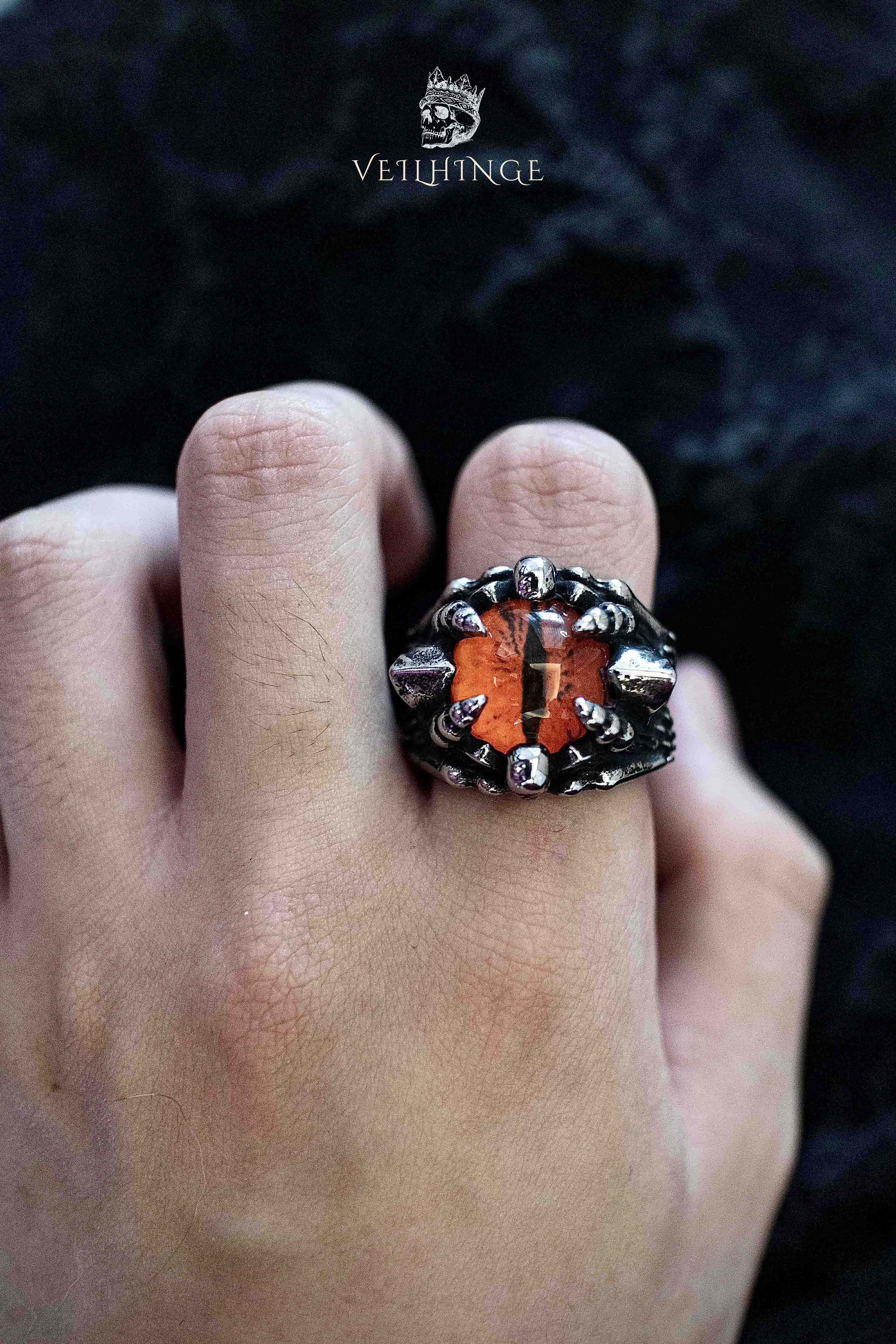 Dragon claw ring gripping an orange faceted stone, with cloaked skull figures on each shoulder, worn on a hand against a black background