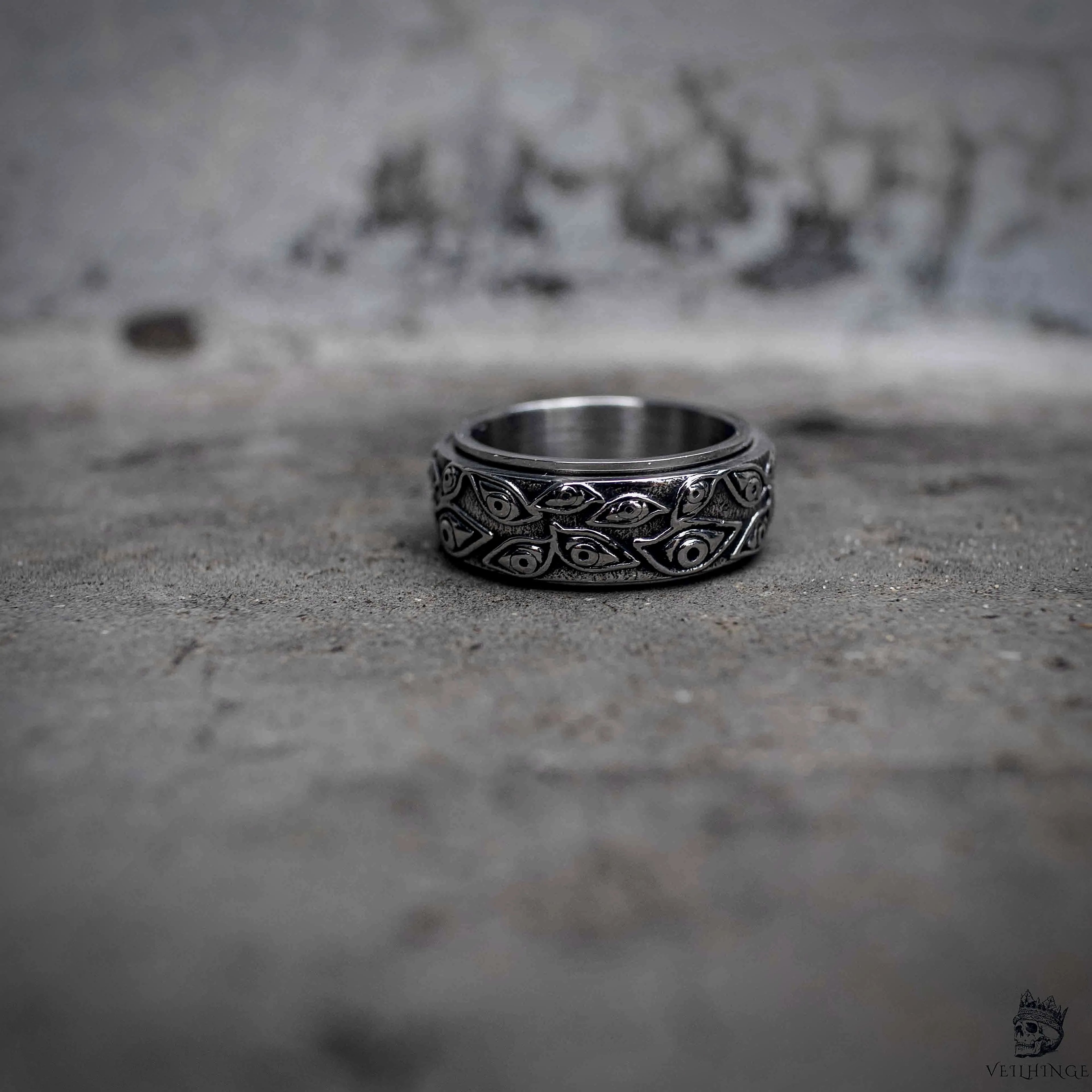 Evil eye spinning ring flat lay showing oxidized outer rotating band with eye engraving and polished inner band, dark aesthetic stainless steel fidget ring by Veilhinge