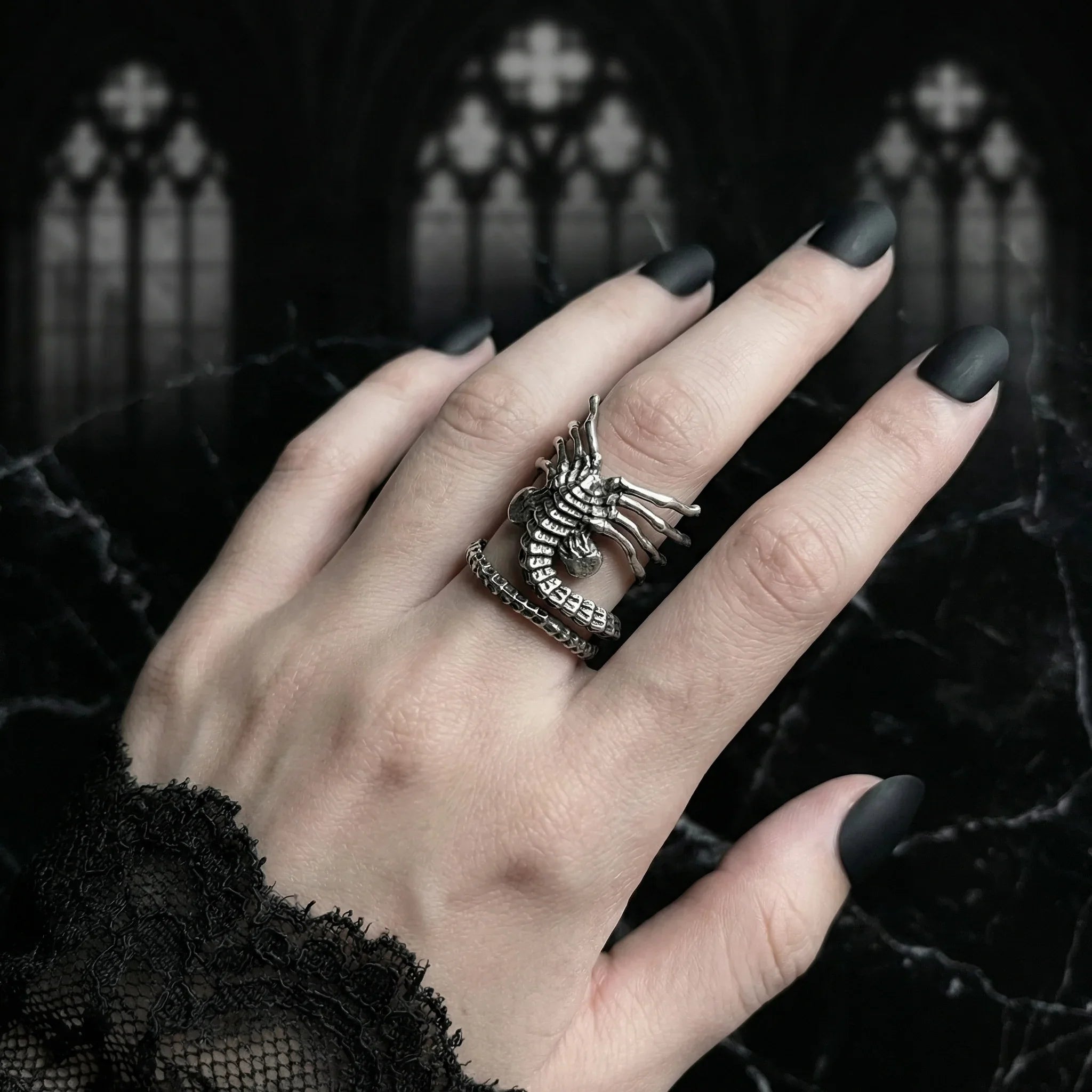 Elegant pale hand with black matte nails wearing the adjustable Facehugger Xenomorph ring against a dark gothic cathedral window background, black lace sleeve