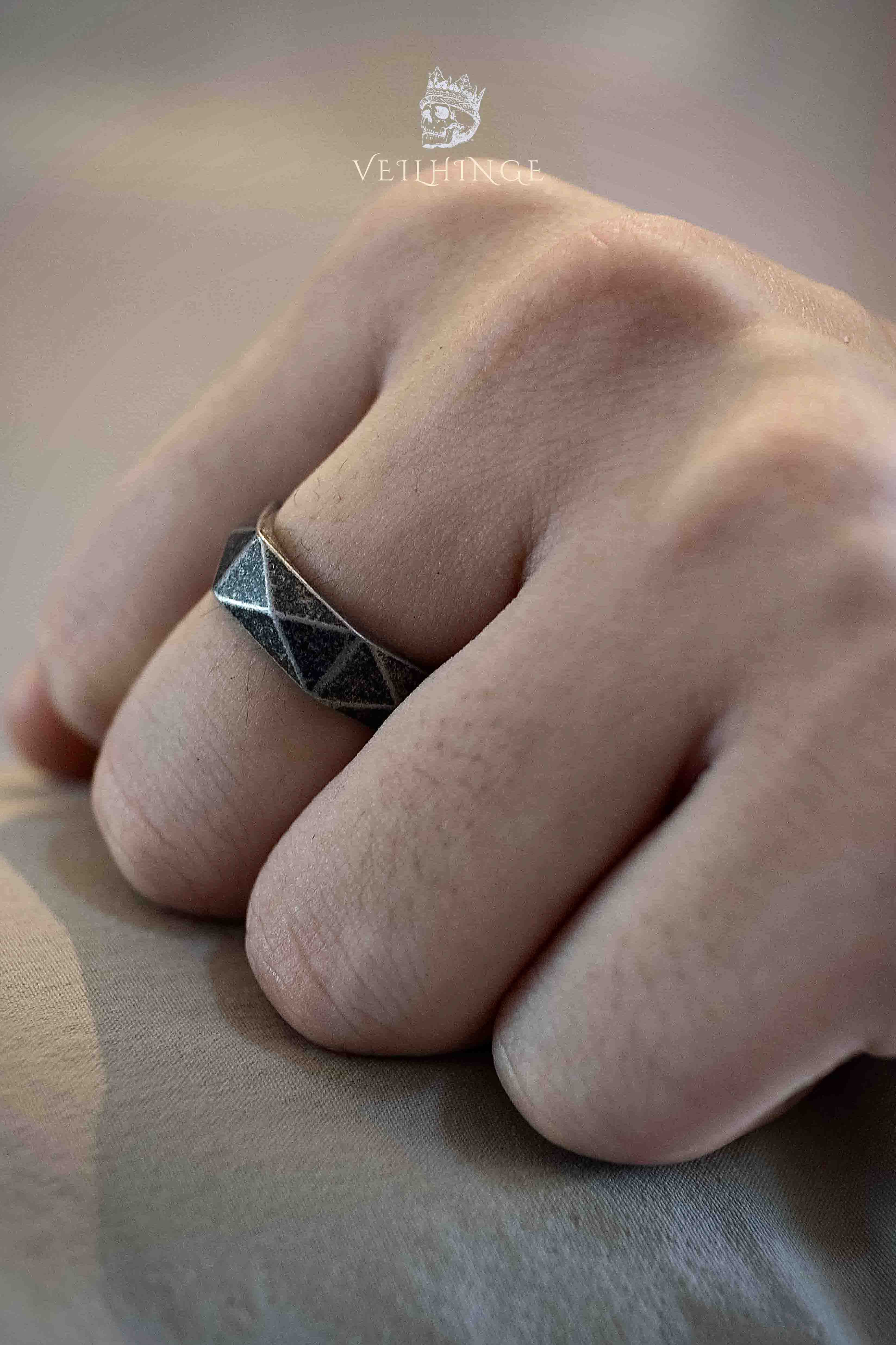 Wide band ring with geometric diamond-cut faceted panels worn on a clenched fist, hammered texture inside each angled section, dark oxidized silver with strong light contrast