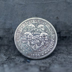 Close-up of a coin engraved with four skulls surrounding an anchor with a two-line Carpe Diem banner, radiant rays spreading from the anchor top, E Pluribus Unum border, dated 1896