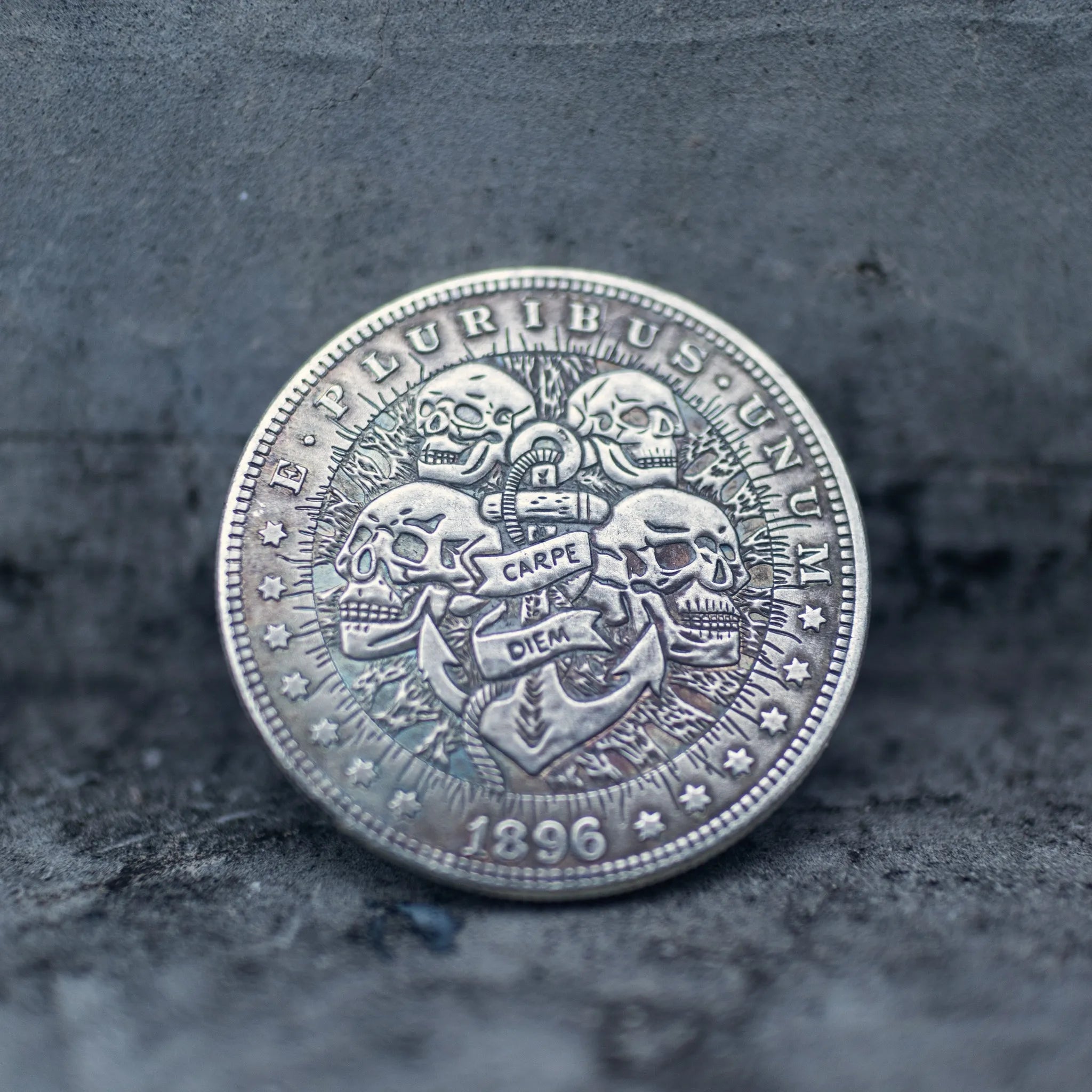 Close-up of a coin engraved with four skulls surrounding an anchor with a two-line Carpe Diem banner, radiant rays spreading from the anchor top, E Pluribus Unum border, dated 1896