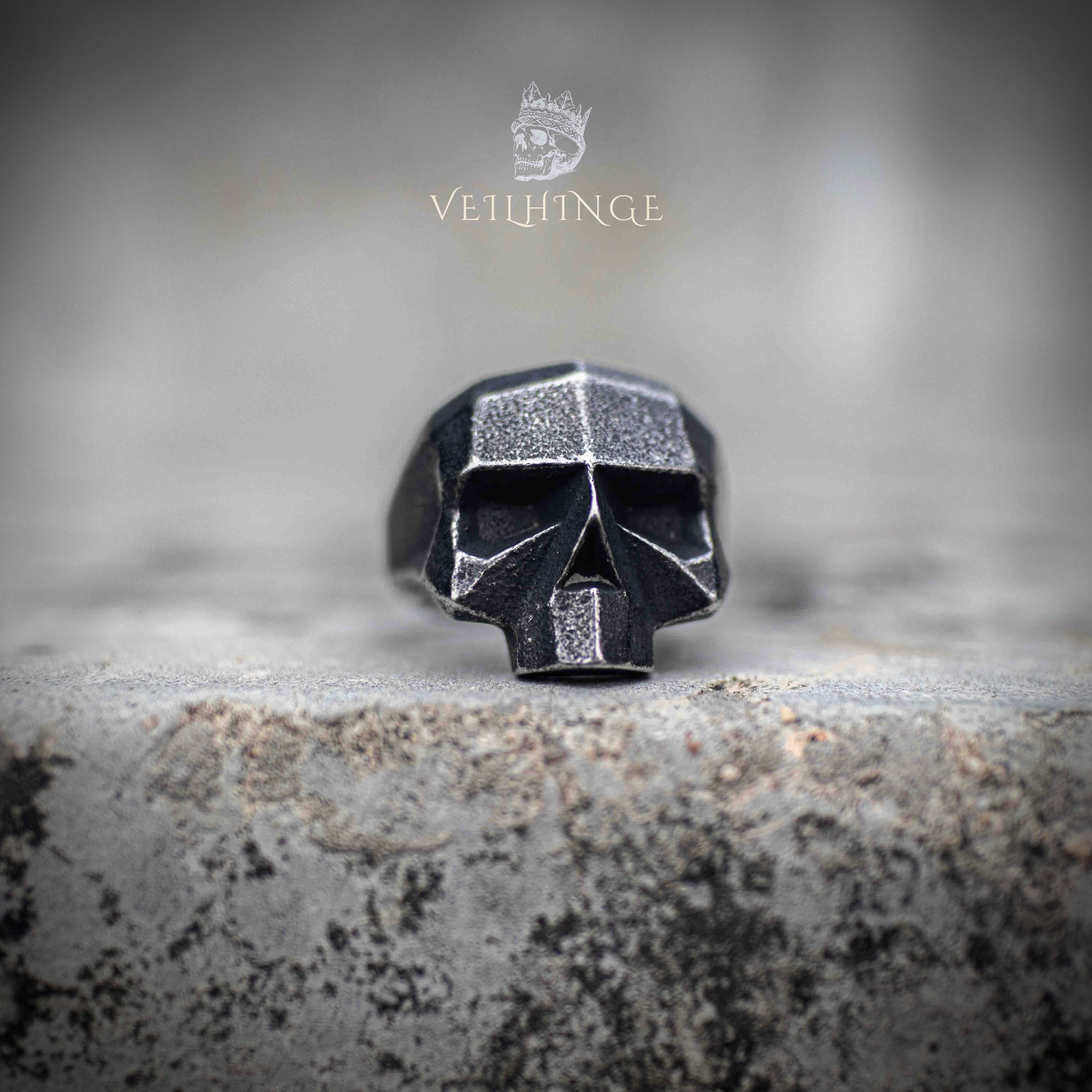 Side profile of geometric skull ring showing oxidized exterior and brushed steel inner band — angular low-poly construction on stone surface Main image